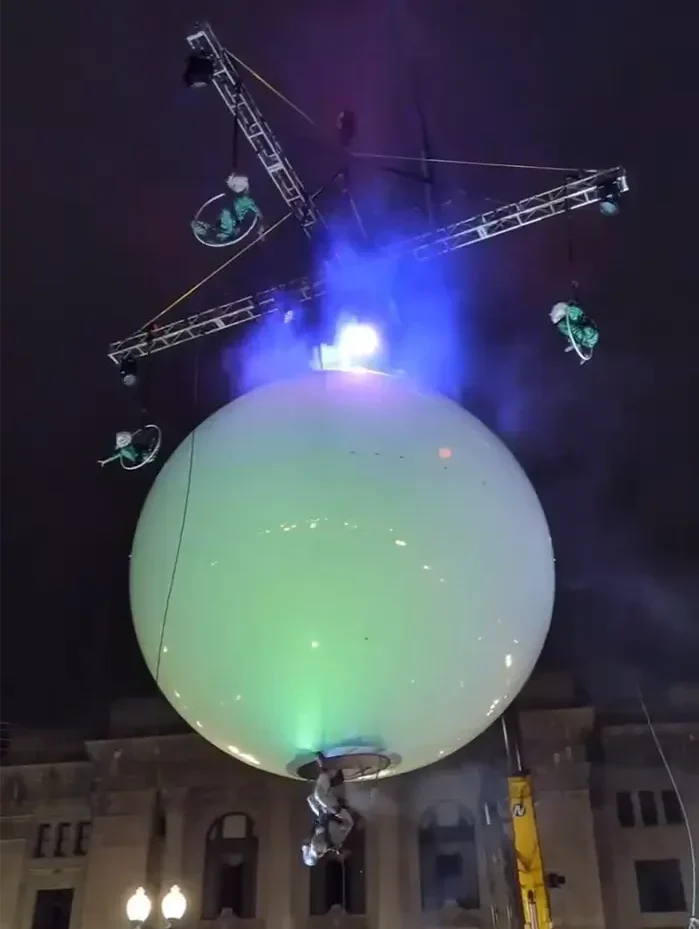 Illuminated Vertical eVenti sphere suspended in the air with aerial performers attached to a cross-shaped truss above